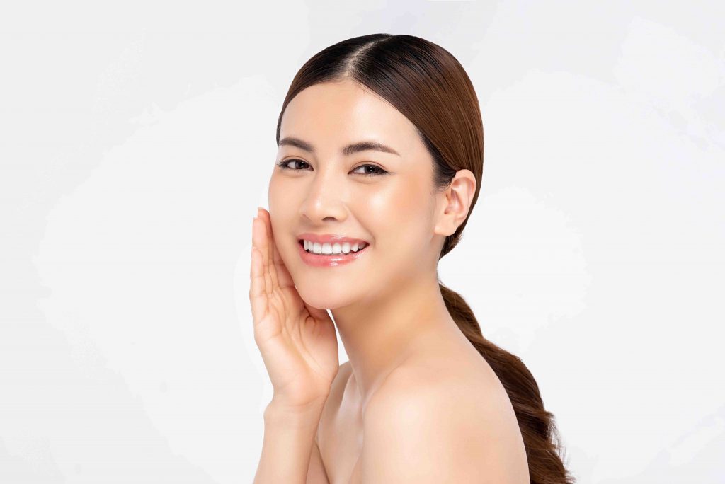 Mulher asiática sorridente com mão no rosto do lado esquerdo. Cabelos longos, usa cabelos amarrados. 