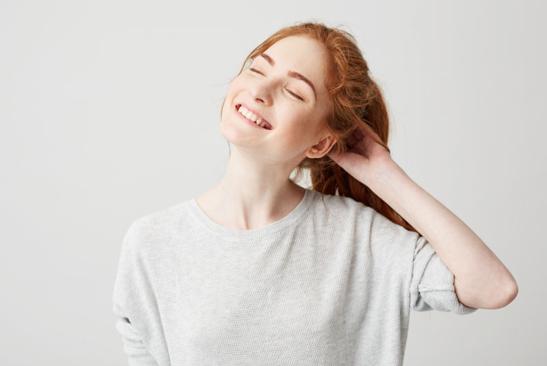 Imagem de uma mulher branca, ruiva, de cabelo preso, sorrindo com os olhos fechados e a cabeça inclinada para o lado esquerdo da imagem. Ela usa uma camiseta cinza e está com a mão esquerda na nuca. O fundo da imagem é cinza, um pouco mais escuro do que a sua blusa.